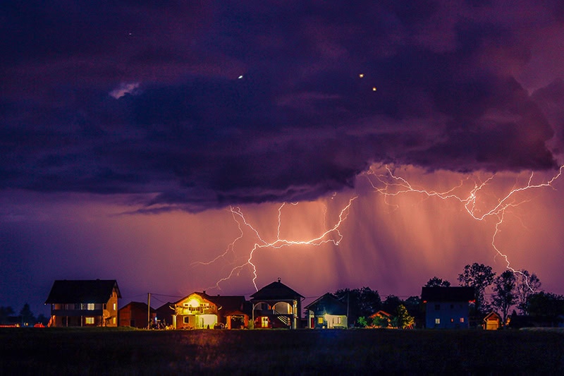 A thunderstorm rolling in at sunset with bolts of lightening striking behind a neighborhood.