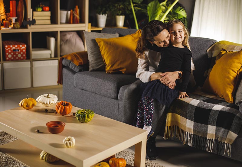 A mother and young daughter embracing on a sofa in a warmly lit living room decorated for autumn and Halloween with small decorative pumpkins on the coffee table.