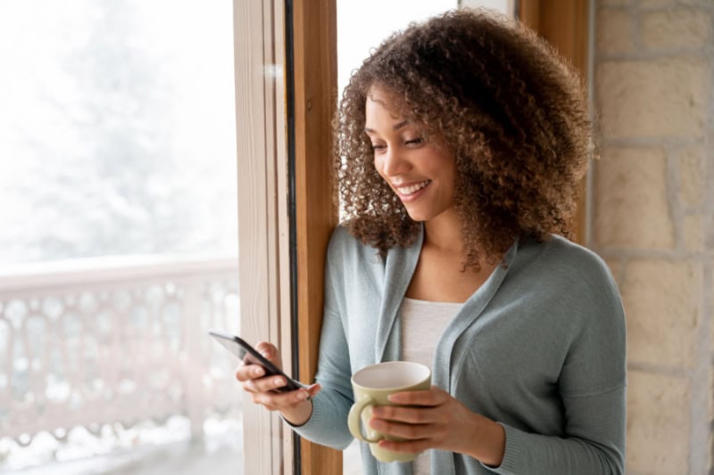 Woman standing at the window in her home smiling at her cell phone.
